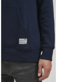 Dunkelblaues Sweatshirt mit Kängurutasche, gerippten Bündchen und Saum sowie einem Stoffetikett mit Pflegehinweisen. Weiches, hochwertiges Material.
