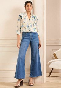 Blusa floreale con maniche a volant, fondo crema, abbinata a jeans a gamba larga blu. Il modello indossa sandali con tacco color cammello. Tessuti morbidi e texture.
