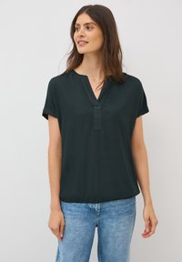 Blouse courte à manches courtes de couleur vert foncé avec un décolleté en V, fabriquée dans un tissu lisse. Le bas présente un léger froncé pour un ajustement décontracté. Portée avec un jean bleu clair.