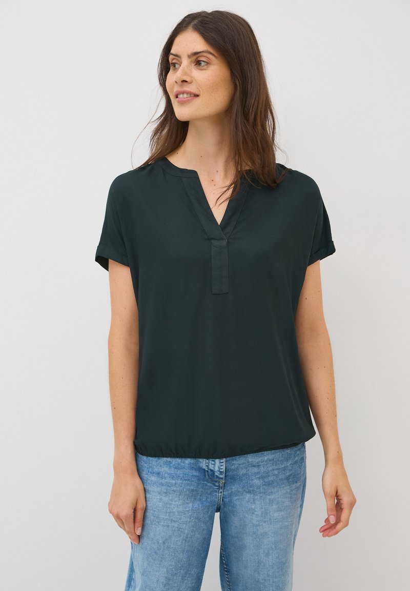 Blouse courte à manches courtes de couleur vert foncé avec un décolleté en V, fabriquée dans un tissu lisse. Le bas présente un léger froncé pour un ajustement décontracté. Portée avec un jean bleu clair.
