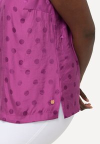 Paarse zijden blouse met een subtiel stippenpatroon, voorzien van een zijzak en een kleine contrasterende knoop aan de onderrand.