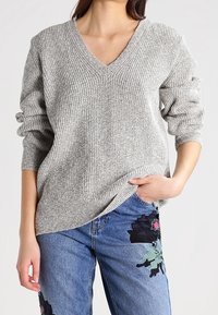 Grau gerippter Pullover mit V-Ausschnitt und langen Ärmeln, kombiniert mit blauen Jeans, die florale Stickerei auf dem linken Oberschenkel haben.