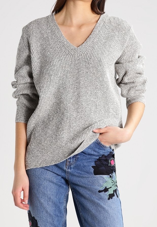 Grau gerippter Pullover mit V-Ausschnitt und langen Ärmeln, kombiniert mit blauen Jeans, die florale Stickerei auf dem linken Oberschenkel haben.