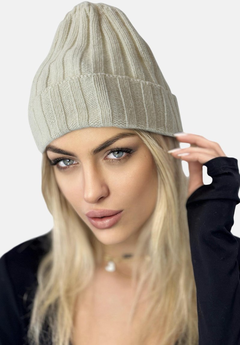 Beanie lavorato a maglia di un beige chiaro con un motivo a coste verticali, texture morbida e design a punta, indossato da una persona con capelli lunghi e biondi.