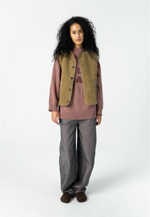 Kvinde med langt krøllet hår iført en brun, fluffy vest over en mauve trøje, grå vidde bukser og mørkebrune sko, stående mod en hvid baggrund.