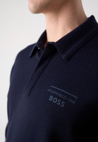 Homme portant un polo bleu marine avec le logo « Porsche BOSS » sur la poitrine gauche, vue rapprochée du col et du haut du torse.