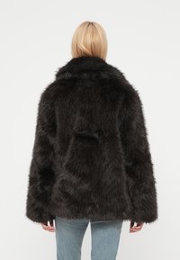 Μαύρο μπουφάν από ψεύτικη γούνα με φαρδύ γιακά και χαλαρή εφαρμογή. Υφή επιφάνειας και μακριά μανίκια, συνδυασμένο με ανοιχτό μπλε τζιν. Εμφάνιση από πίσω.