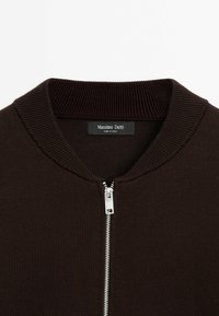 Maglione lavorato a maglia marrone con chiusura a zip, caratterizzato da un colletto e polsini a coste. L'etichetta riporta "Massimo Dutti", indicando l'origine del marchio.