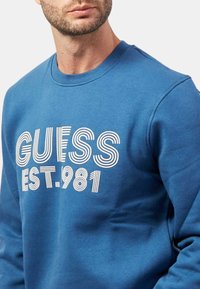 Felpa blu in cotone misto con colletto a girocollo a coste e la scritta "GUESS EST. 1981" in carattere bianco a righe sul davanti.