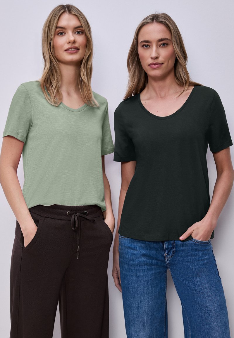 Deux femmes se tiennent côte à côte, portant des chemises à manches courtes décontractées, l'une vert clair avec un pantalon noir, l'autre vert foncé avec un jean bleu.
