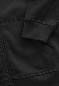 Haut à capuche zippé noir avec des bords côtelés, texture lisse et poche latérale. Met en avant un design minimaliste avec des détails de couture robustes.