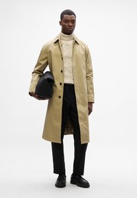 Beige waterdichte trenchcoat met zwarte knopen, gelaagd over een crèmekleurige kabeltrui en zwarte broeken, met een zwarte tas.