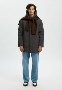 Cappotto impermeabile grigio con cappuccio, indossato sopra un maglione chiaro, abbinato a jeans azzurri e scarpe bianco e nere. Sciarpa marrone annodata attorno al collo.