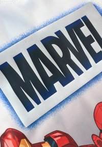 Fehér anyag, amelyen egy kiemelkedő, fekete színű "MARVEL" logó található ezüst háttérrel és kék színátmenetes díszítéssel. Karakterábrázolások is vannak rajta.