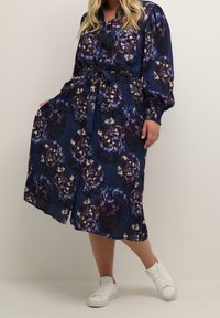 Navyblauwe jurk met een groot bloemenpatroon in tinten paars en crème, met een knoopsluiting aan de voorkant, een ceintuur in de taille en lange, opgeblazen mouwen.