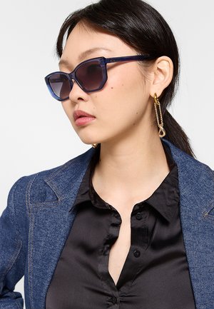 Donna con capelli scuri indossa occhiali da sole scuri oversize, orecchini a catena dorati, una camicia nera e una giacca di jeans blu, guarda verso sinistra.