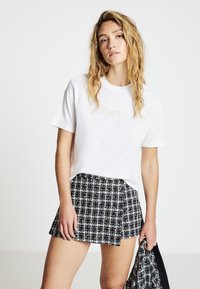 Weißes Baumwoll-T-Shirt mit dem Schriftzug "élégance" in goldener Stickerei, kombiniert mit einem schwarz-weiß karierten Minirock mit goldenen Akzenten.