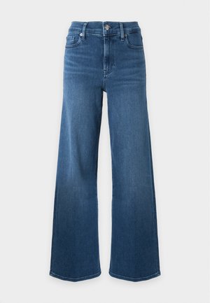 Femme debout, main sur la hanche, portant un haut blanc sans manches, un jean bleu taille haute à jambes larges, une ceinture noire et des sandales à talons noirs.