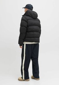 Sort pufferjakke med hætte og en quiltet design. Bæres med marineblå bukser med en hvid side-stribe og beige støvler.