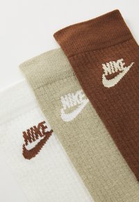 Trois paires de chaussettes en blanc, beige et marron. Fabriquées en tissu tricoté avec des bords côtelés, chacune arborant le logo Nike dans des couleurs contrastées.