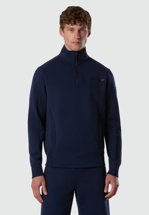 Junger Mann mit lockigem Haar, der einen marineblauen Pullover mit Viertelreißverschluss und passende Hosen trägt, stehend vor einem schlichten, hellen Hintergrund.