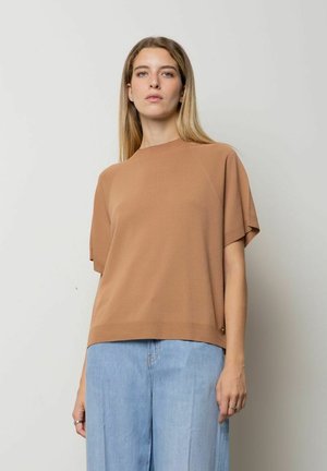 Donna con lunghi capelli biondi che indossa un maglione marrone corto e largo e jeans azzurri a gamba larga, in piedi davanti a uno sfondo semplice.