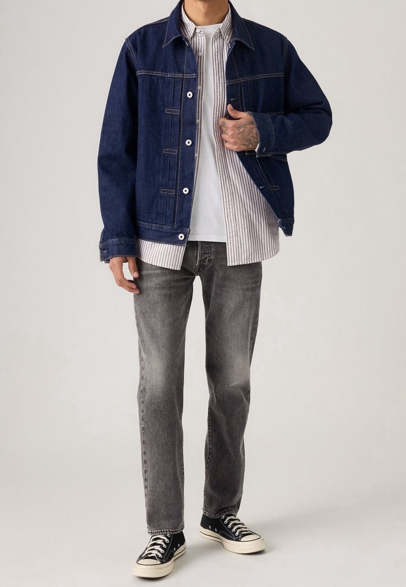 Homme portant une veste en denim bleu foncé par-dessus une chemise rayée, un t-shirt blanc, un jean gris et des baskets montantes noires, debout devant un fond uni.