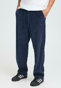 Homme portant un pantalon en velours côtelé bleu marine et des baskets noires avec des rayures blanches, les mains dans les poches, stand sur un fond clair.