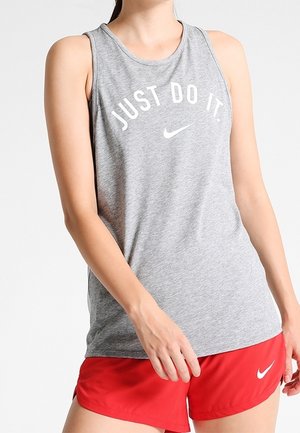 Femme portant un haut sans manches gris avec le texte "JUST DO IT." et un short rouge Nike, debout avec une main près de sa cuisse.