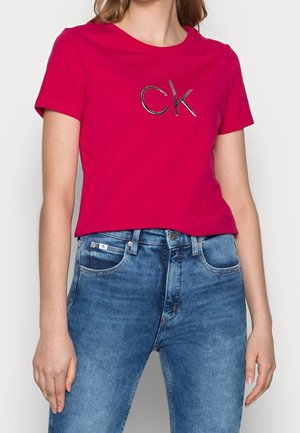 Kobieta ubrana w jasnoczerwoną koszulkę z krótkim rękawem z czarnym logo "ck" oraz wysokim stanem niebieskich dżinsów, stojąca na jednolitym tle.