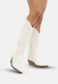 Botas de vaquero de cuero blanco con punta afilada, diseño acolchado y tacón de madera marrón. Cremallera lateral para ponerlas y quitarla fácilmente; detallado de costura intrincada.