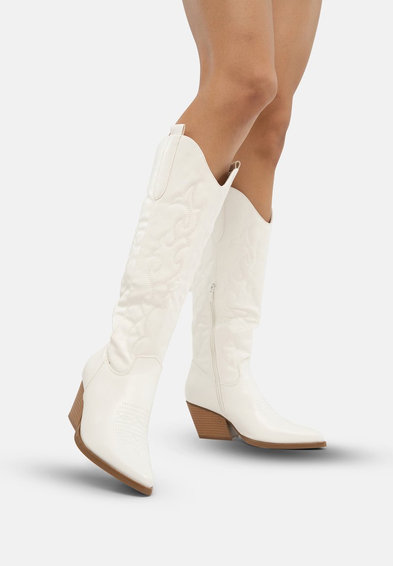 Botas de vaquero de cuero blanco con punta afilada, diseño acolchado y tacón de madera marrón. Cremallera lateral para ponerlas y quitarla fácilmente; detallado de costura intrincada.