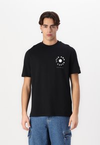 Μαύρη βαμβακερή μπλούζα T-shirt με κοντά μανίκια, στρογγυλή λαιμόκοψη, λογότυπο στο μπροστινό μέρος του στήθους σε λευκό χρώμα, συνδυασμένη με γαλάζιες τζιν cargo σορτς.