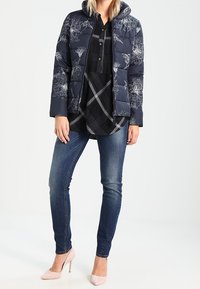 Donna che indossa un piumino floreale blu navy, una camicia a quadri nera, jeans skinny blu e tacchi alti rosa chiaro, in piedi contro uno sfondo semplice.
