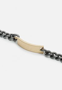 Pulseira metálica com elos de corrente pretos e um centro de detalhe em ouro liso e curvado. Apresenta um hardware resistente e um design minimalista.