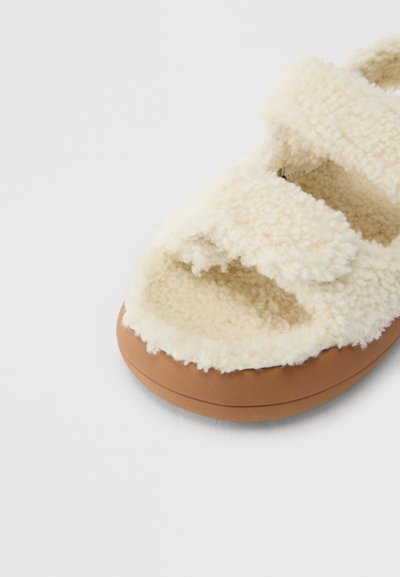 Sandale à enfiler en fausse fourrure blanche avec trois brides réglables, dotée d'une semelle en caoutchouc marron, d'une texture douce et d'une semelle intérieure rembourrée.