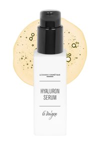 Hyaluronserum in einer rechteckigen weißen Flasche mit schwarzem Pumpspender. Das Etikett enthält den Produktnamen und die Marke. Gelbe Flüssigkeitsunterlage.