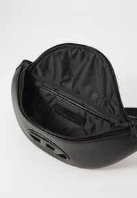 Schwarze runde Tasche mit glattem Äußeren und auffälligem Logo. Das Innere ist mit schwarzem Stoff ausgekleidet, der ein sich wiederholendes Logomuster aufweist. Mit Reißverschluss.