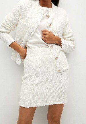 Kvinde iført en hvid tekstureret jakke med knapper over en ensfarvet hvid t-shirt og en matchende tekstureret mininederdel.