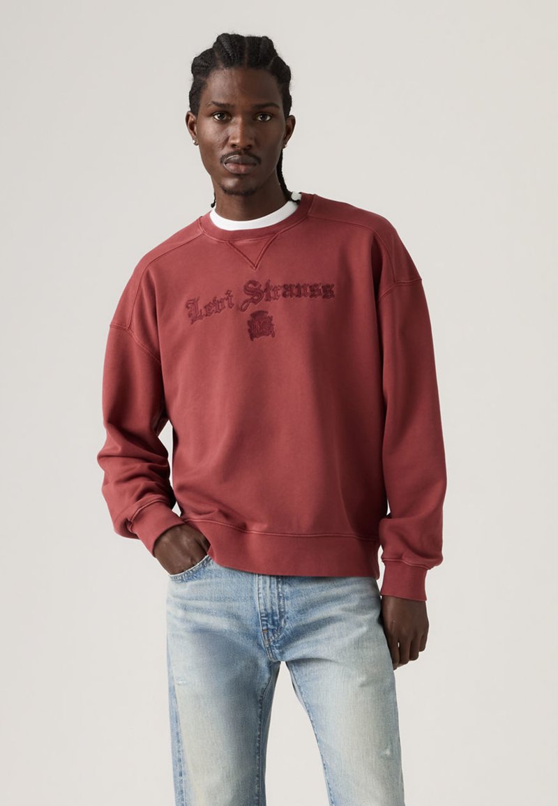 Φούτερ τούβλινο κόκκινο από βαμβάκι με λαιμόκοψη crew και κεντημένο λογότυπο "Levi Strauss". Συνδυάζεται με ανοιχτόχρωμα μπλε τζιν.