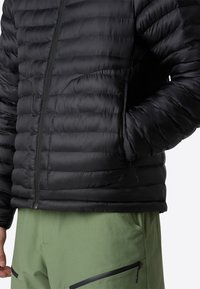 Chaqueta de plumas negra con patrón acolchado horizontal, cierre de cremallera y bolsillos laterales; llevada sobre pantalones verdes con acentos negros.