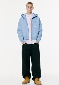 Haut-de-vêtement bleu clair à capuche, zippé sur le devant et avec logo, superposé à une chemise rayée rose ; associé à un pantalon en denim noir ample et des bottes beiges.