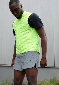 Hombre con chaleco amarillo neón, camiseta negra de manga corta y pantalones cortos grises de Nike, de pie al aire libre contra una pared gris.