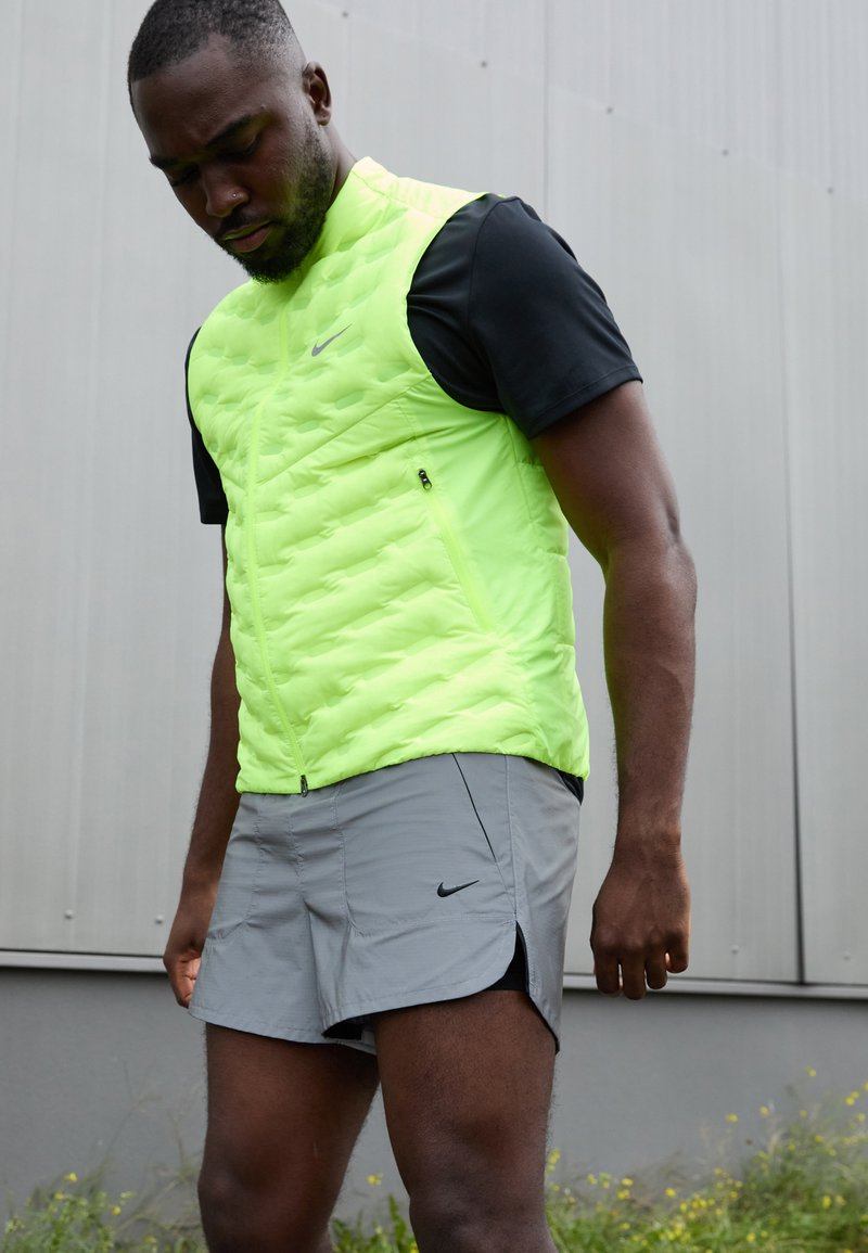 Hombre con chaleco amarillo neón, camiseta negra de manga corta y pantalones cortos grises de Nike, de pie al aire libre contra una pared gris.