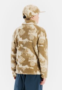 Chaqueta de forro polar con un patrón en tonos marrones y crema, de cuello alto y mangas largas. Una gorra a juego cuenta con una visera plana y una correa ajustable.