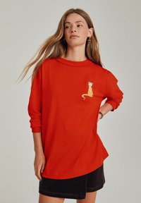Tricou roșu supradimensionat din bumbac, cu un design de leopard brodat în zona stângă a pieptului. Croială lejeră, cu mâneci 3/4.