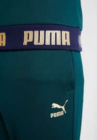 Tyrkysové sportovní kalhoty s texturovaným povrchem, s širokým námořnickým pasem s nápisem "PUMA" ve zlaté barvě a zlatým logem na dolní části nohavice.