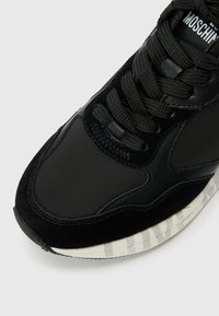 Baskets de sport noires avec un mélange de tissus lisses et de textures en suède. Lacets épais noirs et semelle en caoutchouc rayée avec un détail de marque.
