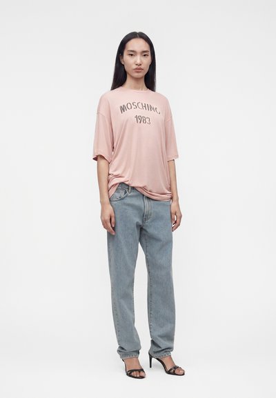 Γυναίκα όρθια, φορεμένη με oversized ανοιχτό ροζ t-shirt Moschino 1983, άνετο ανοιχτό μπλε τζιν και μαύρα πέδιλα με ανοιχτά δάχτυλα και τακούνι, πάνω σε λευκό φόντο.