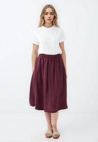 Jupe midi bordeaux avec une taille élastique, fabriquée en tissu léger, elle présente une silhouette fluide et une texture lisse. T-shirt blanc et sandales.
